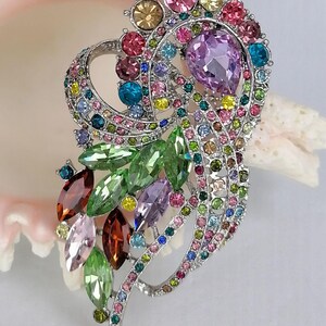 May include: A silver-tone brooch featuring a swirling, openwork design encrusted with multicolored rhinestones. The piece includes marquise-cut green, brown, and pink crystals, along with a prominent pear-shaped lavender stone and small round gems.