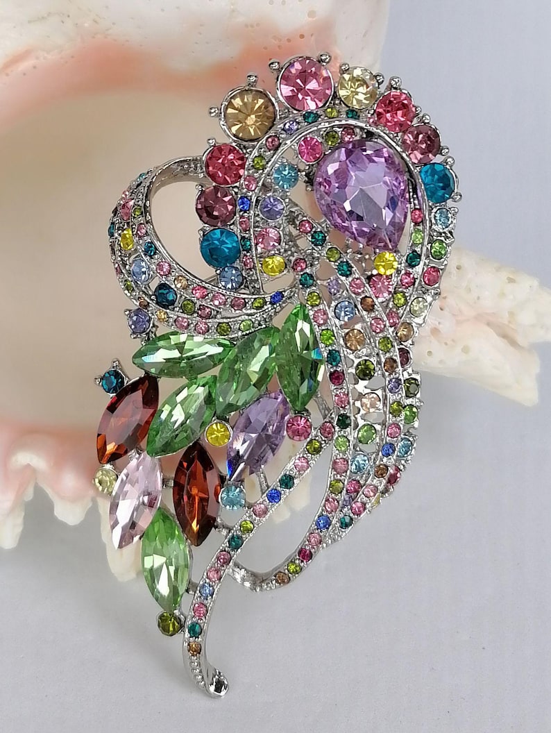 May include: A silver-tone brooch featuring a swirling, openwork design encrusted with multicolored rhinestones. The piece includes marquise-cut green, brown, and pink crystals, along with a prominent pear-shaped lavender stone and small round gems.