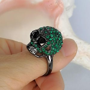Op de afbeelding: Een ring van zwart metaal met een schedelvormig middendeel. De schedel is ingelegd met talloze kleine, levendige groene kristallen die de bovenkant en zijkanten bedekken, terwijl de oogkassen en de neusholte hol en donker blijven.