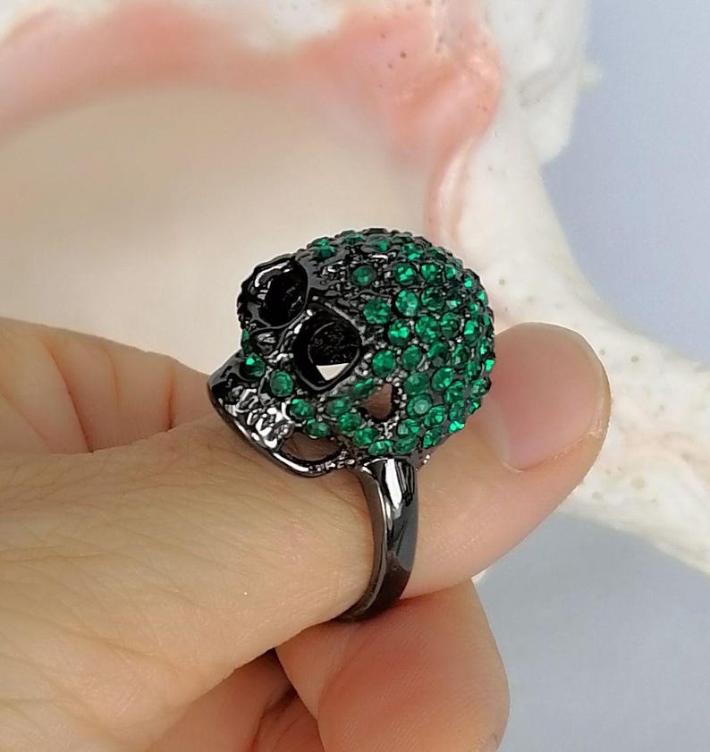 Op de afbeelding: Een ring van zwart metaal met een schedelvormig middendeel. De schedel is ingelegd met talloze kleine, levendige groene kristallen die de bovenkant en zijkanten bedekken, terwijl de oogkassen en de neusholte hol en donker blijven.