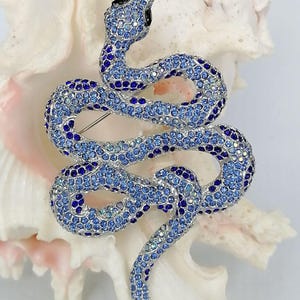 May include: A blue and silver jewelled snake brooch. The snake is coiled, with a black head and a pin on the back. The brooch is covered in small, sparkling crystals. The brooch is set against a white background.