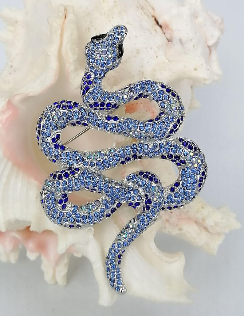 May include: A blue and silver jewelled snake brooch. The snake is coiled, with a black head and a pin on the back. The brooch is covered in small, sparkling crystals. The brooch is set against a white background.