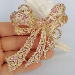 May include: A gold-toned bow brooch, intricately designed with openwork and adorned with pink rhinestones. The brooch has a classic bow shape with flowing tails, perfect for adding a touch of elegance to any outfit.