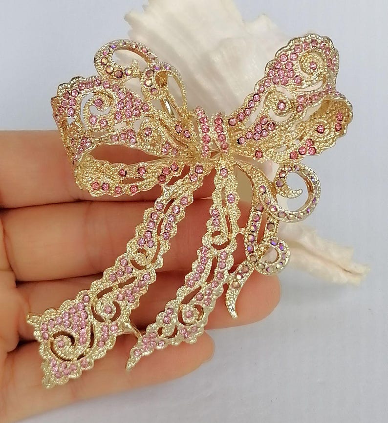 May include: A gold-toned bow brooch, intricately designed with openwork and adorned with pink rhinestones. The brooch has a classic bow shape with flowing tails, perfect for adding a touch of elegance to any outfit.