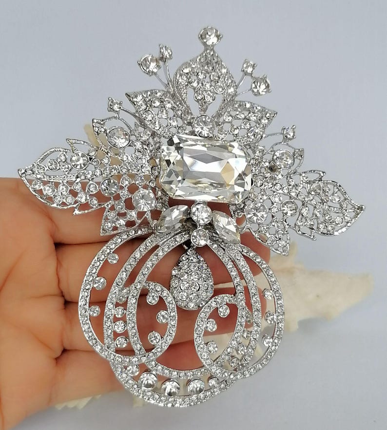 May include: A large silver-toned brooch featuring a prominent rectangular clear crystal at the center, surrounded by intricate filigree metalwork encrusted with numerous small round rhinestones and a central teardrop-shaped crystal pendant.