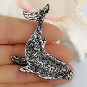 May include: A silver-toned whale brooch with a textured surface. The brooch features a detailed design of a whale with its tail raised. The pin is designed to be attached to clothing or accessories.