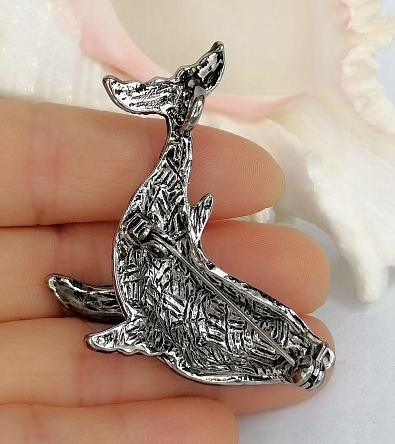 May include: A silver-toned whale brooch with a textured surface. The brooch features a detailed design of a whale with its tail raised. The pin is designed to be attached to clothing or accessories.