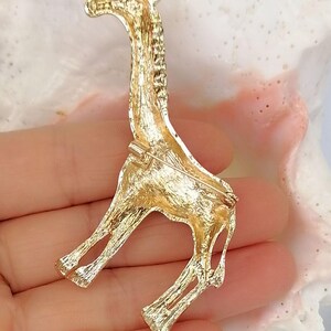 May include: A gold-toned metal brooch shaped like a giraffe, featuring a textured surface finish. The back of the brooch includes a horizontal pin mechanism with a rotating clasp, designed for securing the piece to fabric or other materials.
