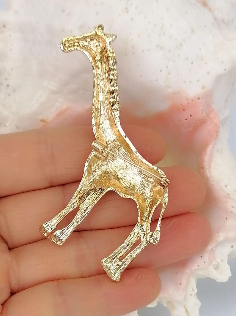 May include: A gold-toned metal brooch shaped like a giraffe, featuring a textured surface finish. The back of the brooch includes a horizontal pin mechanism with a rotating clasp, designed for securing the piece to fabric or other materials.
