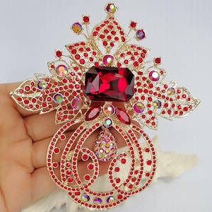 May include: A gold-tone metal brooch featuring a large rectangular deep red central stone, surrounded by numerous small red rhinestones and iridescent circular crystals. The design includes intricate filigree patterns and a teardrop-shaped pendant.