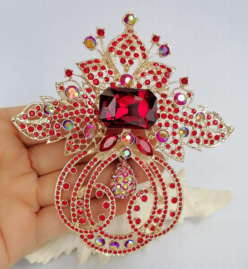 May include: A gold-tone metal brooch featuring a large rectangular deep red central stone, surrounded by numerous small red rhinestones and iridescent circular crystals. The design includes intricate filigree patterns and a teardrop-shaped pendant.