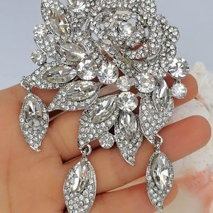 Puede incluir: Un broche plateado con forma de flor y hojas, cubierto totalmente de brillantes piedras de estr&aacute;s transparentes. El dise&ntilde;o presenta una flor central, varias piedras de corte marquesa y tres colgantes en forma de hoja en la parte inferior.