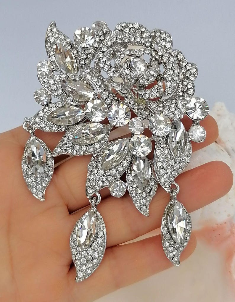 Puede incluir: Un broche plateado con forma de flor y hojas, cubierto totalmente de brillantes piedras de estr&aacute;s transparentes. El dise&ntilde;o presenta una flor central, varias piedras de corte marquesa y tres colgantes en forma de hoja en la parte inferior.