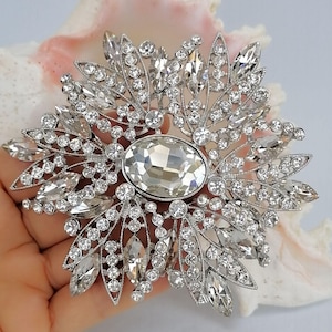 Op de afbeelding: Een zilverkleurige broche met een grote, centrale, ovaal geslepen heldere kristal, omringd door een radiale opstelling van kleinere ronde en marquise-geslepen heldere kristallen, wat een stervormig patroon met filigraan metaaldetails cre&euml;ert.