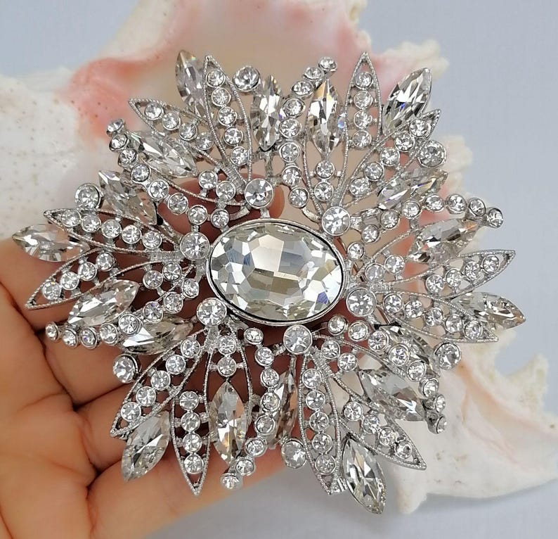 Op de afbeelding: Een zilverkleurige broche met een grote, centrale, ovaal geslepen heldere kristal, omringd door een radiale opstelling van kleinere ronde en marquise-geslepen heldere kristallen, wat een stervormig patroon met filigraan metaaldetails cre&euml;ert.