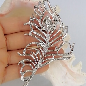May include: A silver-coloured feather brooch with a central, round, clear gemstone. The brooch has an openwork design, with delicate lines and curves that mimic the structure of a feather. The pin is visible on the back.