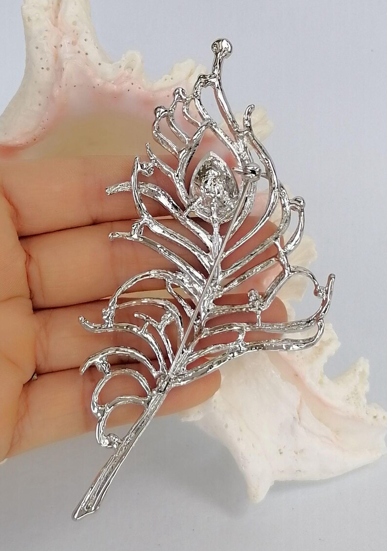 May include: A silver-coloured feather brooch with a central, round, clear gemstone. The brooch has an openwork design, with delicate lines and curves that mimic the structure of a feather. The pin is visible on the back.