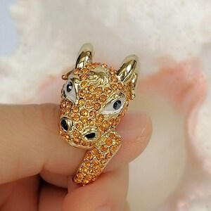 May include: A gold-toned ring featuring a three-dimensional ram head design. The surface of the ram is covered in small, sparkling orange crystals. The ram has smooth, polished gold horns, white enamel eye details with black pupils, and black nostrils.