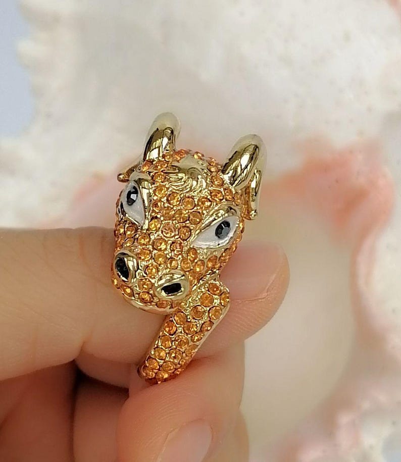 May include: A gold-toned ring featuring a three-dimensional ram head design. The surface of the ram is covered in small, sparkling orange crystals. The ram has smooth, polished gold horns, white enamel eye details with black pupils, and black nostrils.