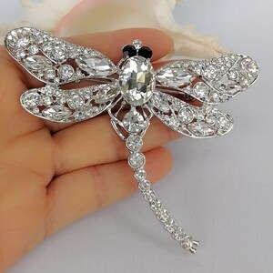 May include: A silver dragonfly brooch, embellished with clear crystals. The brooch has a large oval crystal at its centre, with smaller crystals outlining the wings and body. The dragonfly's head is decorated with black detailing.