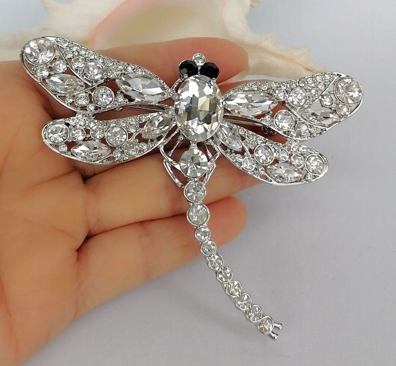May include: A silver dragonfly brooch, embellished with clear crystals. The brooch has a large oval crystal at its centre, with smaller crystals outlining the wings and body. The dragonfly's head is decorated with black detailing.