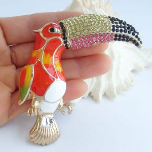 May include: A toucan brooch with a gold-tone metal frame. The bird's body is red, orange, yellow, and white enamel. The beak is encrusted with yellow, pink, and black rhinestones. The brooch is a decorative accessory.