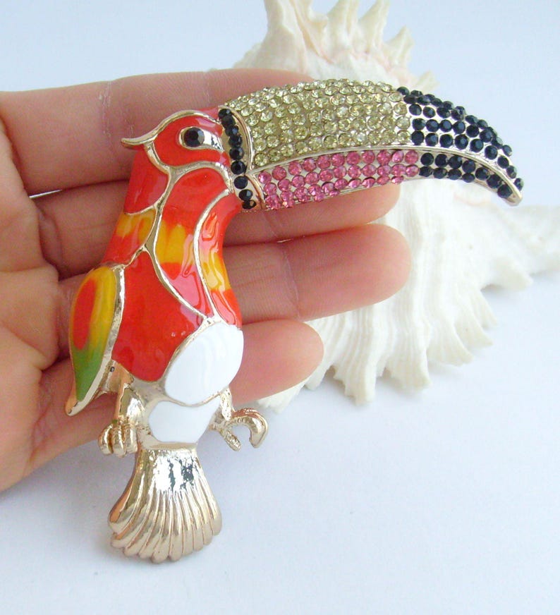 May include: A toucan brooch with a gold-tone metal frame. The bird's body is red, orange, yellow, and white enamel. The beak is encrusted with yellow, pink, and black rhinestones. The brooch is a decorative accessory.