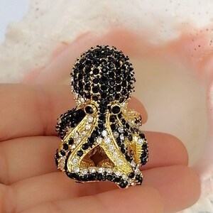Peut inclure: Une broche en forme de pieuvre dor&eacute;e, orn&eacute;e de nombreuses petites pierres scintillantes noires et blanches transparentes sur la t&ecirc;te et les tentacules. La pieuvre poss&egrave;de deux yeux en cristal noir et une base en m&eacute;tal dor&eacute; textur&eacute;.