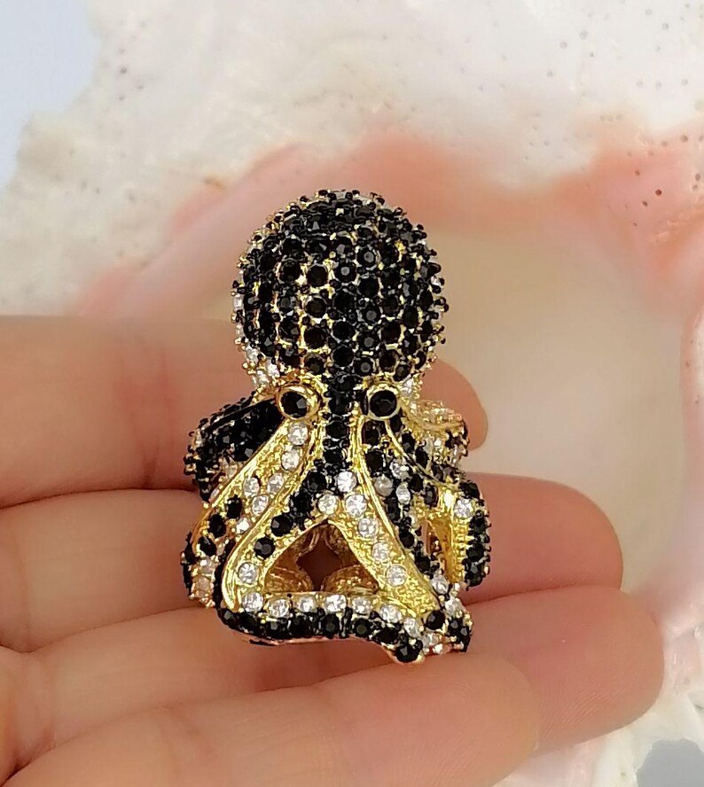 Peut inclure: Une broche en forme de pieuvre dor&eacute;e, orn&eacute;e de nombreuses petites pierres scintillantes noires et blanches transparentes sur la t&ecirc;te et les tentacules. La pieuvre poss&egrave;de deux yeux en cristal noir et une base en m&eacute;tal dor&eacute; textur&eacute;.