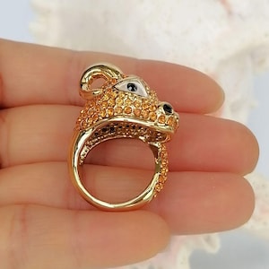 May include: A gold-toned ring shaped like a stylised animal head, featuring a smooth metal band and a surface covered in small, sparkling orange crystals. The animal design includes a white triangular eye detail and a black nose accent.