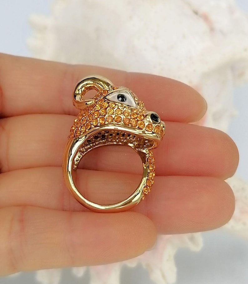 May include: A gold-toned ring shaped like a stylised animal head, featuring a smooth metal band and a surface covered in small, sparkling orange crystals. The animal design includes a white triangular eye detail and a black nose accent.