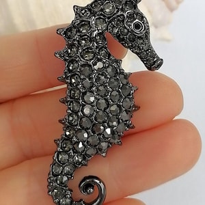 May include: A seahorse-shaped brooch in dark gray, encrusted with small, sparkling crystals. The brooch has a detailed design, including a curled tail and a textured body. The seahorse is a unique accessory for jackets, bags, or scarves.
