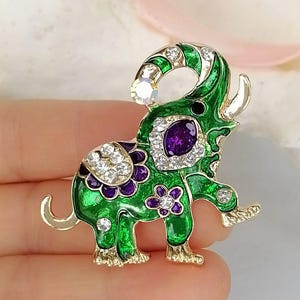 May include: An elephant-shaped brooch featuring vibrant green enamel, gold-tone metal edges, and clear crystal accents. The design includes a prominent purple oval stone on the side, purple floral details, and a single crystal at the tip of the trunk.