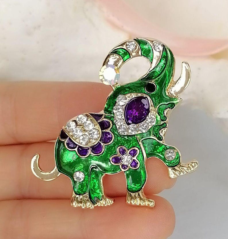 May include: An elephant-shaped brooch featuring vibrant green enamel, gold-tone metal edges, and clear crystal accents. The design includes a prominent purple oval stone on the side, purple floral details, and a single crystal at the tip of the trunk.