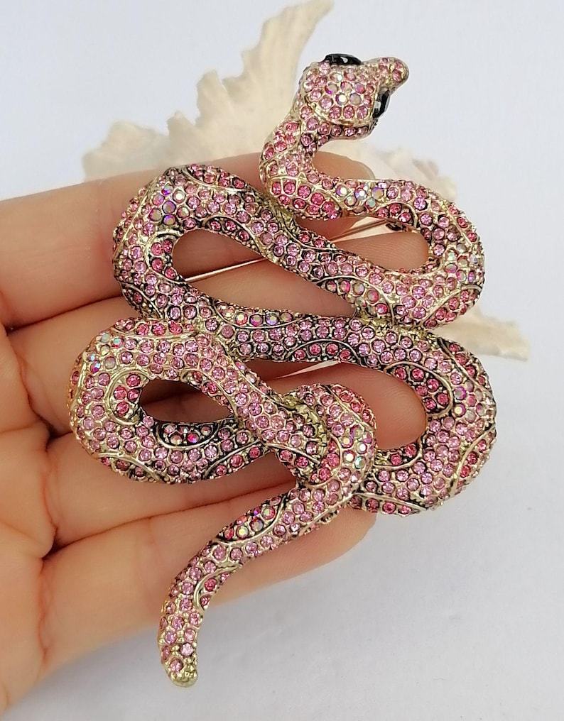 May include: A pink and gold snake brooch. The brooch is encrusted with pink rhinestones and has a black rhinestone eye. The snake is coiled and has a gold-coloured metal base. The brooch is a decorative accessory.