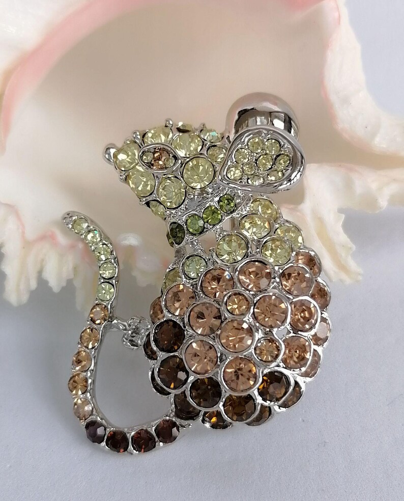 May include: A silver-toned mouse brooch encrusted with a variety of sparkling crystals in shades of yellow, green, and brown. The brooch has a detailed design with a rounded body and a long, curved tail. The mouse's head features a small, round, silver-colored detail.
