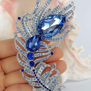May include: A silver-toned feather brooch encrusted with numerous light blue and dark blue crystals. The design features two prominent pear-shaped blue crystals at the center, surrounded by smaller pav&eacute;-set stones arranged in a plume pattern.