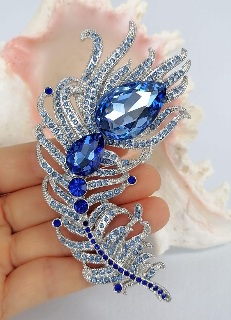 May include: A silver-toned feather brooch encrusted with numerous light blue and dark blue crystals. The design features two prominent pear-shaped blue crystals at the center, surrounded by smaller pav&eacute;-set stones arranged in a plume pattern.