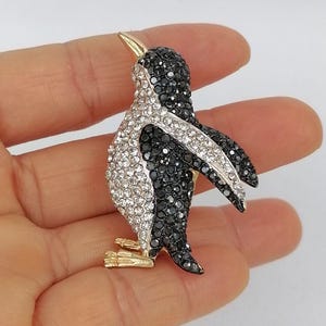 May include: A penguin brooch with a gold beak and feet. The penguin's body is covered in black and clear crystals, creating a sparkling effect. The brooch is a decorative accessory.