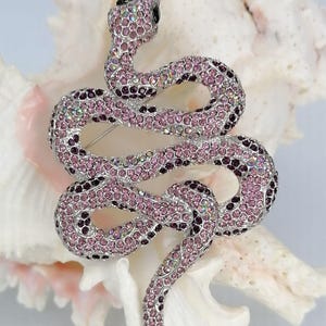 May include: A decorative snake brooch, encrusted with pink and purple crystals. The snake design features a coiled body and a black eye. The brooch is set on a white, textured background.