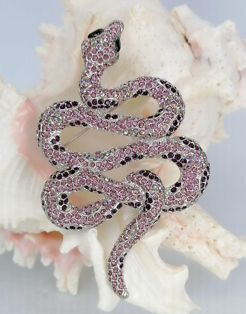May include: A decorative snake brooch, encrusted with pink and purple crystals. The snake design features a coiled body and a black eye. The brooch is set on a white, textured background.