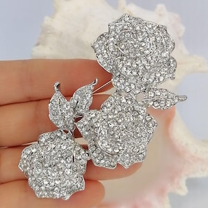 May include: A silver-toned brooch featuring three rose blossoms and two leaves, entirely encrusted with clear, sparkling rhinestones. The metal structure is polished silver, and the design displays a cluster of floral elements with intricate petal detailing.