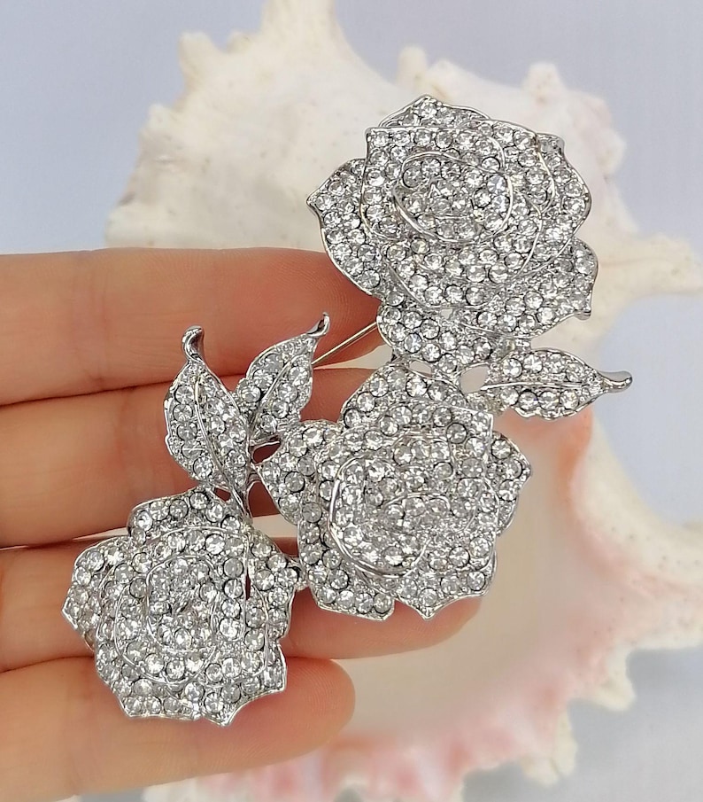 May include: A silver-toned brooch featuring three rose blossoms and two leaves, entirely encrusted with clear, sparkling rhinestones. The metal structure is polished silver, and the design displays a cluster of floral elements with intricate petal detailing.
