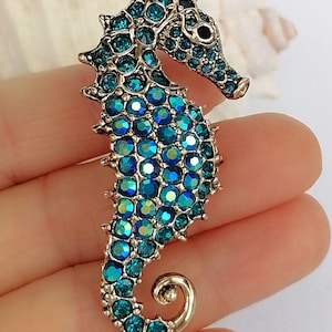 May include: A seahorse-shaped brooch with a silver-tone metal base. The seahorse is encrusted with teal and blue crystals. The brooch has a curved tail and a detailed head with a small eye.