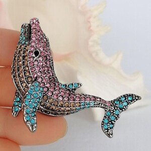 May include: A bejeweled whale brooch with a silver-tone finish. The whale is encrusted with pink, brown, and turquoise rhinestones. The brooch is a decorative accessory.