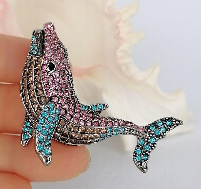 May include: A bejeweled whale brooch with a silver-tone finish. The whale is encrusted with pink, brown, and turquoise rhinestones. The brooch is a decorative accessory.