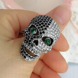 May include: A skull-shaped ring featuring a split design covered in small, sparkling crystals. One side of the skull is encrusted with black crystals, while the other side features clear white crystals. Two vibrant green crystals serve as eyes.