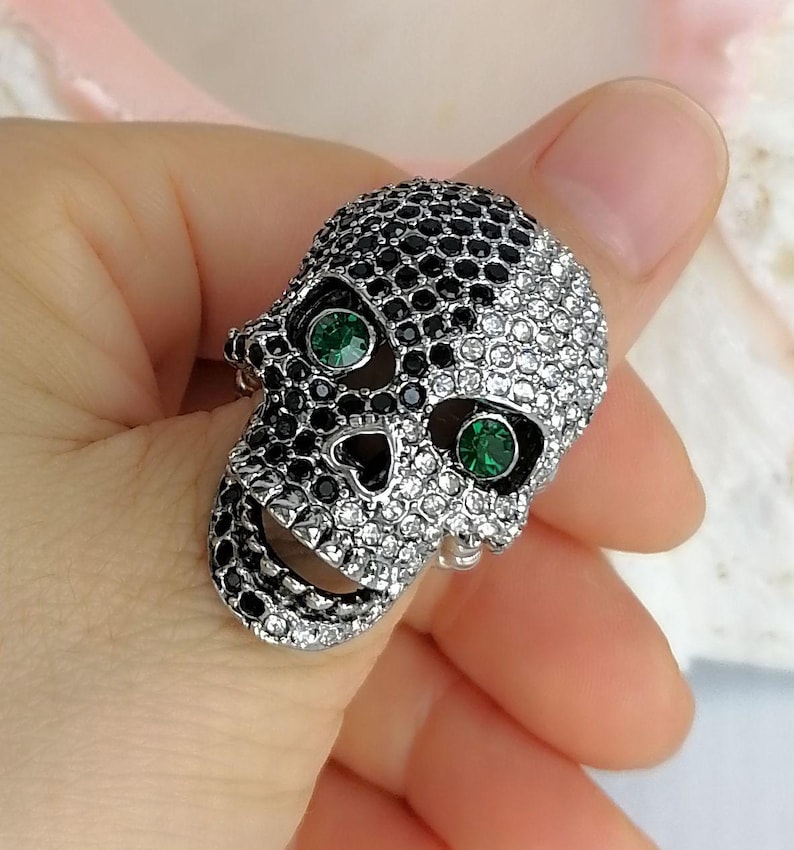 May include: A skull-shaped ring featuring a split design covered in small, sparkling crystals. One side of the skull is encrusted with black crystals, while the other side features clear white crystals. Two vibrant green crystals serve as eyes.
