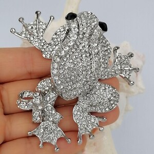 K&ouml;nnte beinhalten: Silberfarbene Froschbrosche, besetzt mit klaren Kristallen. Der Frosch hat schwarze Augen und detaillierte F&uuml;&szlig;e. Diese dekorative Brosche ist dazu bestimmt, Kleidung oder Accessoires zum Funkeln zu bringen.