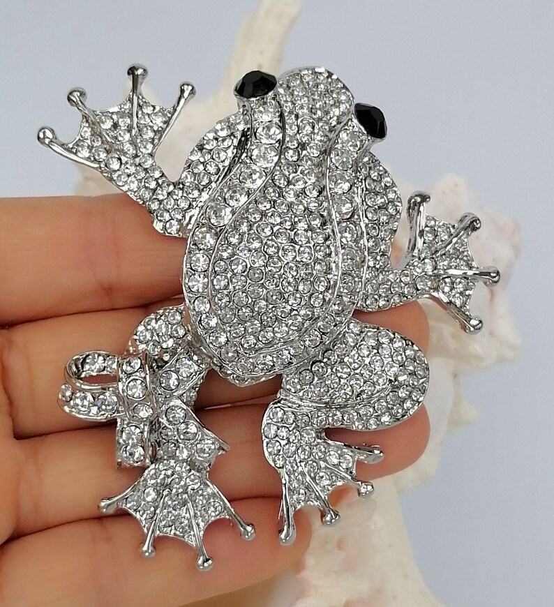 K&ouml;nnte beinhalten: Silberfarbene Froschbrosche, besetzt mit klaren Kristallen. Der Frosch hat schwarze Augen und detaillierte F&uuml;&szlig;e. Diese dekorative Brosche ist dazu bestimmt, Kleidung oder Accessoires zum Funkeln zu bringen.